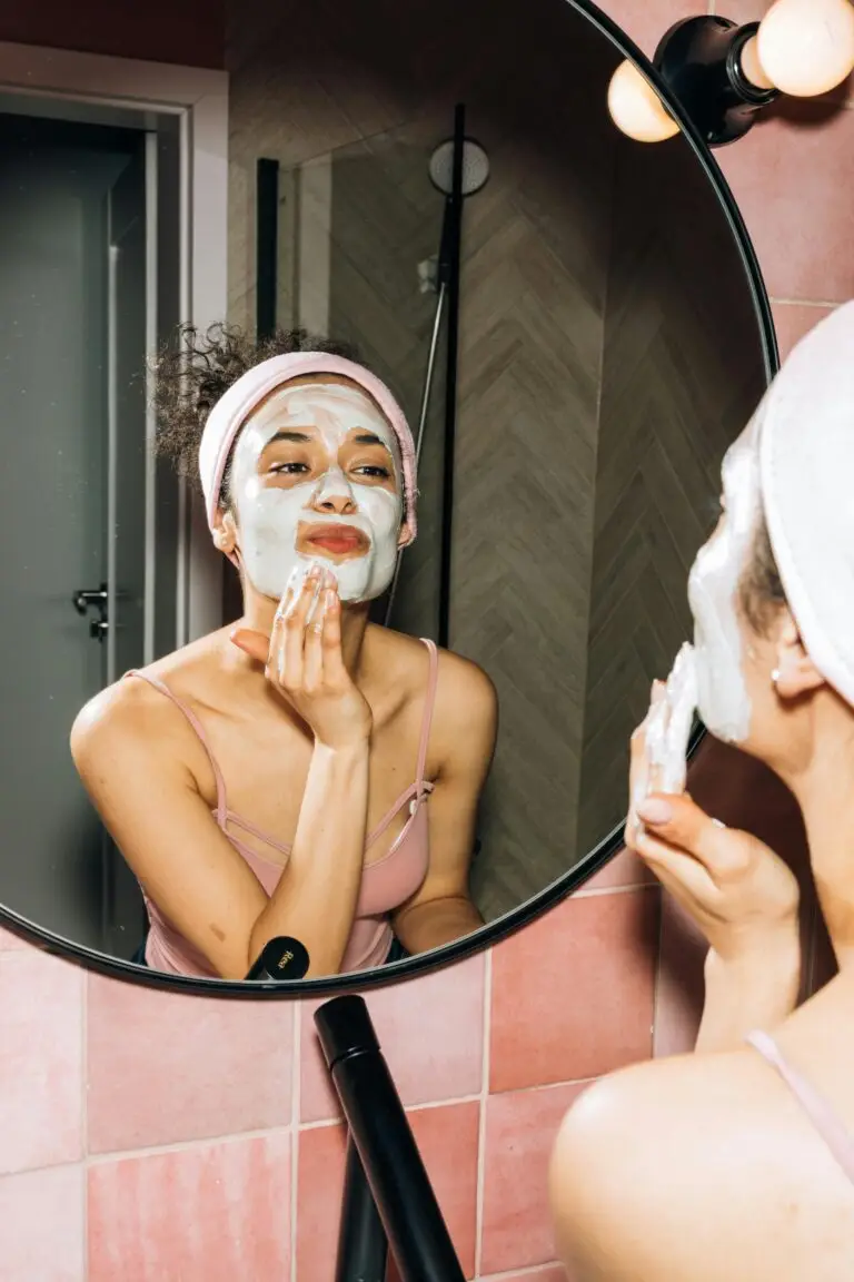 A young woman smiling while applying a facial mask during her home skincare routine.